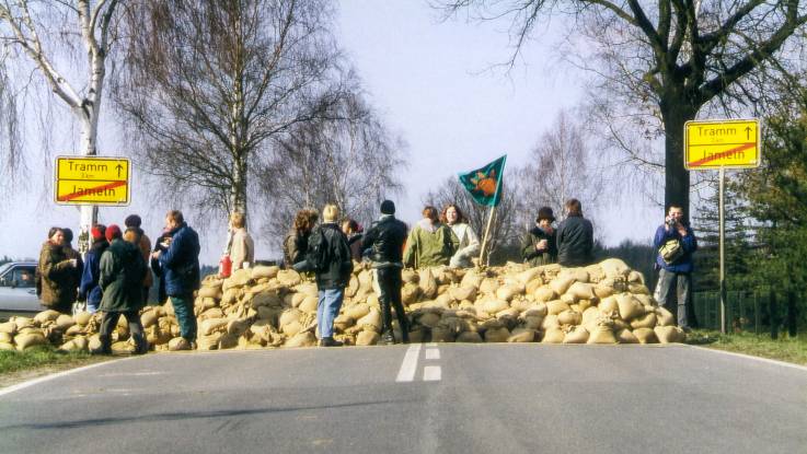 Auf einer Landstraße ist eine etwa ein Meter hohe Straßensperre mit Sandsäcken eingerichtet. Es stehen einige Leute darum herum.