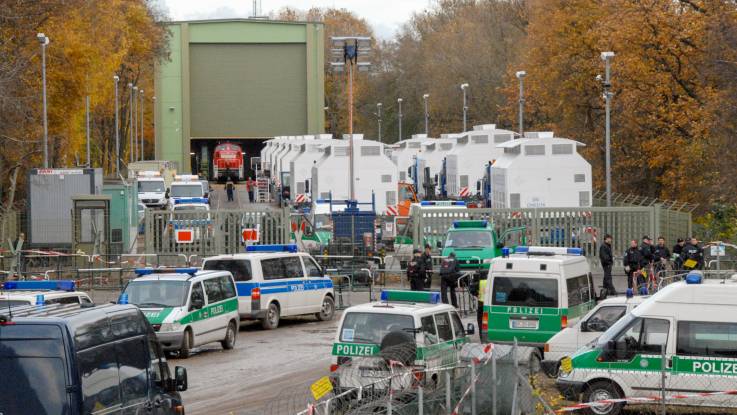 In einem herbstlichen Wald steht eine große Halle, davor mindestens 10  weiße Spezialbehälter auf Bahnwaggons. Das Gelände ist abgesperrt, davor stehen zahlreiche Polizeiwagen. 