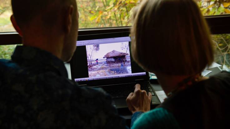 Die Köpfe eines Mannes und einer Frau beugen sich über einen Laptopbildschirm, auf dem das Bild einer zerstörten Hütte zu sehen ist. 