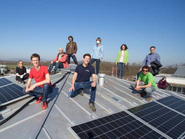 Eine kleine Gruppe von Menschen, auf einem flachen Dach stehend oder kniend, um sie sind PV-Module montiert