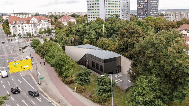 Luftaufnahme: Neben einer mehrspurigen Straße befindet sich ein eingeschossiges kubisches Gebäude mit einem geschwungenen polierten Dach, umgeben von Bäumen.