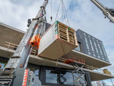 Ein riesiger Kran hievt ein aus Holz gefertigtes Gebäudemodul in das im Rohbau befindliche Obergeschoss aus Beton.