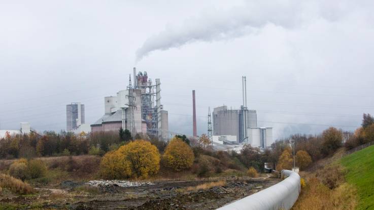 In herbstlicher Landschaft steht in der Ferne ein großes Industriewerk mit hohen rauchenden Schloten, davor führt ein Förderband durch eine trostlose Brache.