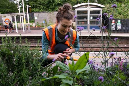 Eine junge Frau in orangefarbener Warnweste gießt Pflanzen etwas oberhalb eines Bahnsteiges der Londoner U-Bahn.