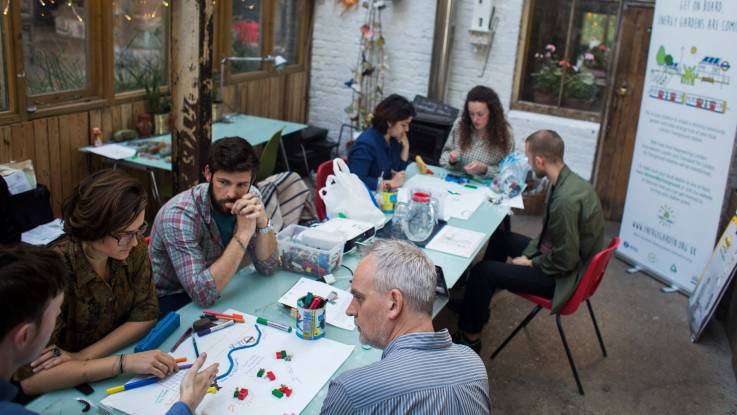 Seven men and women sit around a table filled with plans and creative materials, engrossed in conversation.