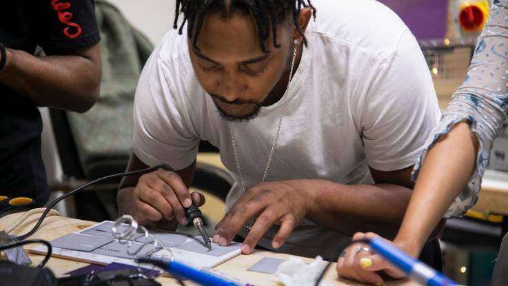 A young man with dark skin and dreadlocks solders small solar panels together.