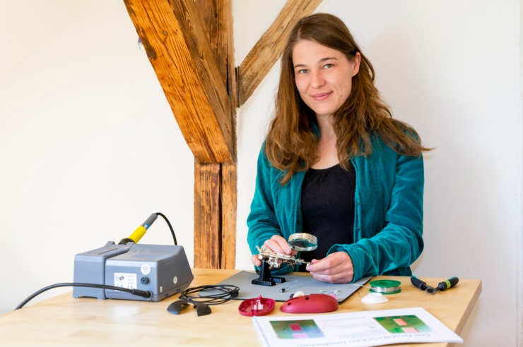 Eine junge Frau hält eine Lötkolben in der Hand, arbeitet an kleinen Platine und schaut offen in die Kamera.