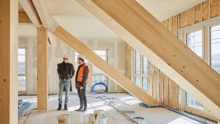 Zwei Männer stehen in einem lichtdurchfluteten Bürogeschoass, zwei massive Holzträger erstrecken sich quer durch den Raum.