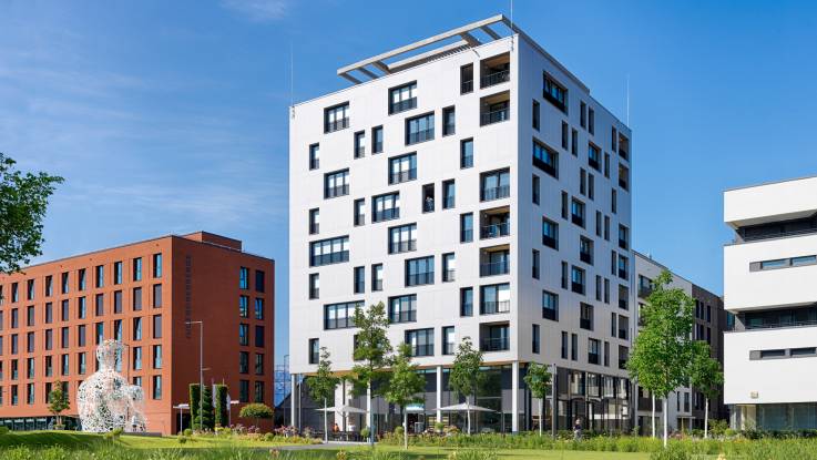 Ein kubisches Gebäude mit nahezu quadratischem Grundriss, dessen weiß schimmernde Fassade von zahlreichen Fensteröffnungen durchbrochen wird.