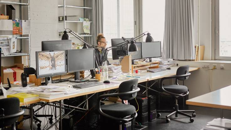 Ein junger Mann mit Brille sitzt an einem von vielen Bürotischen, auf denen große Computerdisplays und Tastaturen zu sehen sind.