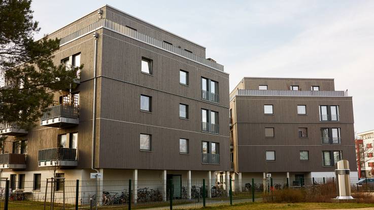 Zwei versetzt zueinander angeordnete kubische Bauten mit graubrauner Fassade; am vorderen der Gebäude sind Balkons zu erkennen.