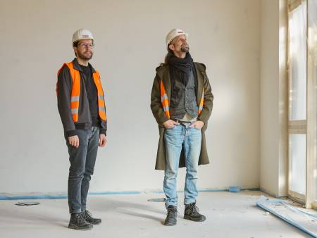 Zwei Männer in Baustellenmontur stehen im Rohbau eines Hauses an einer Fensterfront.