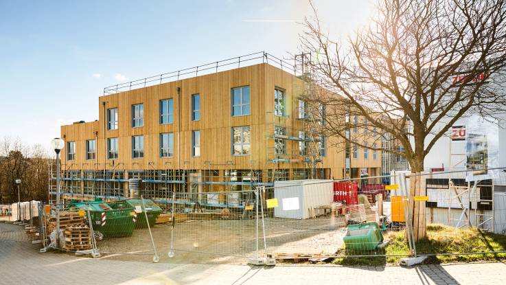 Hinter einem Bauzaun befindet sich ein dreigeschossiges Gebäude mit einer Holzfassade.