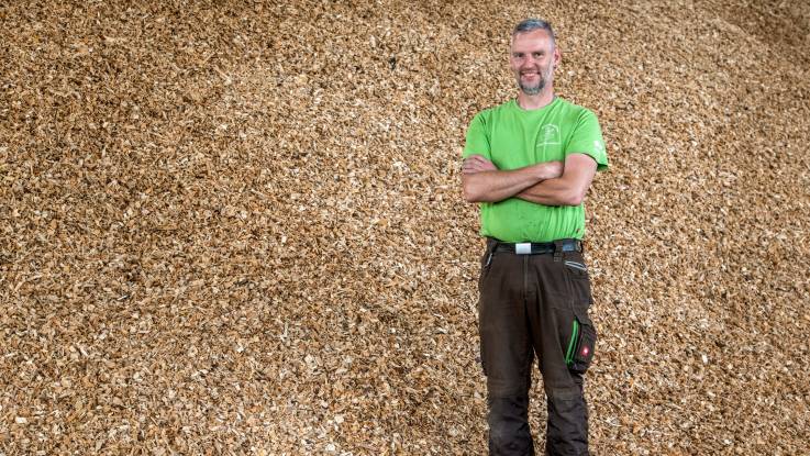 Ein junger Mann in Arbeitshose und einem grünen T-Shirt steht inmitten eines Haufens mit Holzhaeckseln.