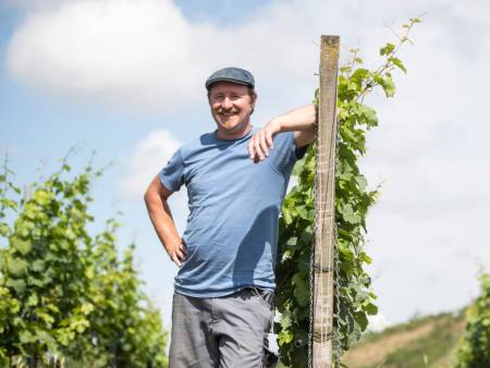 Ein kerniger, sehr tatkräftig wirkender Mann lehnt an einem Pfosten im Weinberg und lächelt in die Kamera.