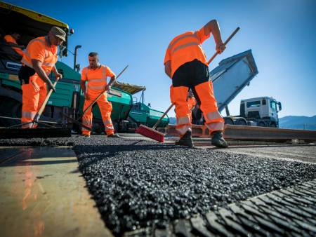 Drei Strassenbauarbeiter stehen vor einer Asphaltiermaschine und verteilen frischen Asphalt mit großen Harken und Besen.