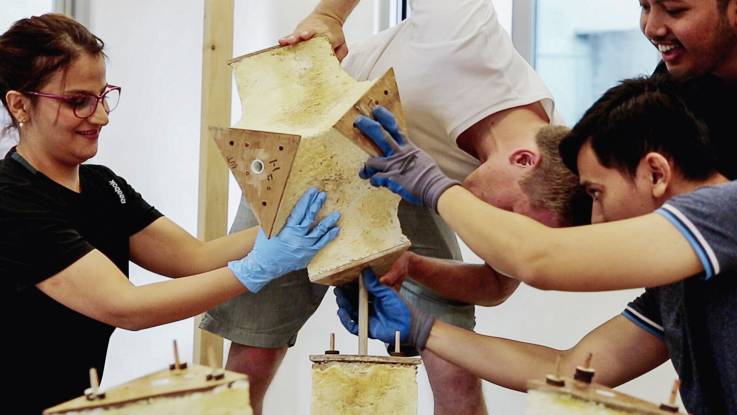 Sechs Hände dreier junger Menschen halten ein Bauelement aus Myzelwerkstoff, das auf ein anderes aufgesteckt wird.