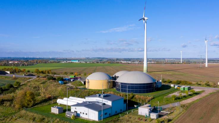 Luftaufnahme: Inmitten der Landschaft stehen drei Windräder, daneben drei Speicher mit kuppelförmigen Dächern sowie weitere Gebäude; auch drei große zigarrenförmige Tanks sind zu erkennen.