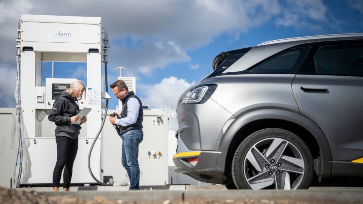 Vor einer Wasserstofftankstelle stehen eine Frau und ein Mann; sie trägt ein Klembrett mit sich, während er den Tankschlauch in der Hand hat. Davor steht ein silberfarbenes Auto.