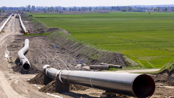 Riesige, noch unverbundene Teilstücke einer Pipeline liegen in einer flachen Baugrube, die von Agrarflächen umgeben ist.