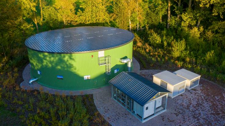 Blick auf einen grünen, kreisrunden Speichertank an einem Waldrand, neben dem einige weitere kleine Zweckbauten angeordnet sind