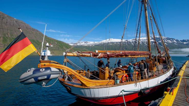 Ein Holzsegelschiff mit deutscher Beflaggung liegt an Land vertäut  in dunklem Wasser, eine karge winterliche Landschaft im Hintergrund.