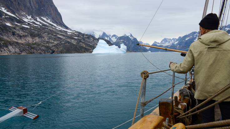 Foto vom Boot aus zeigt eine Person, die an einer Art Angel einen Behälter  im Wasser mitzieht. Im Hintergrund eine karge Berglandschaft mit Schnee.