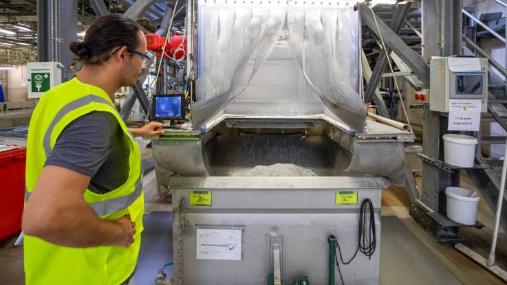 An einer Maschine steht ein Arbeiter mit Warnweste. Die Maschine hat eine offene Einfüllluke, in der ein Haufen grauen pulvrigen Materials zu sehen ist.