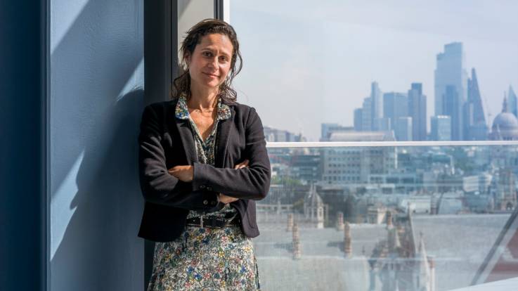 Eine junge Frau lehnt vor einem großen Fenster, hinter dem Hochhäuser zu sehen sind.