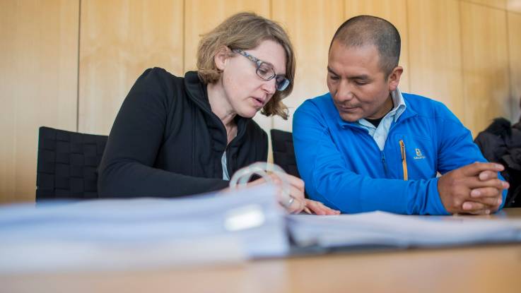 Eine Frau mit aschblondem Haar und Brille sitzt zusammen mit einem Mann südamerikanischen Mann über Akten vertieft an einem Tisch.