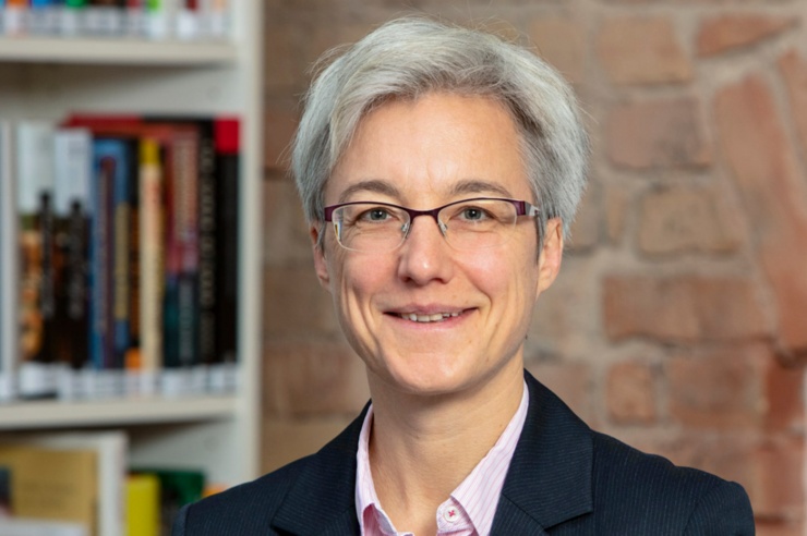 Portrait einer Frau mit kurzen grauen Haaren vor einer Backsteinwand.