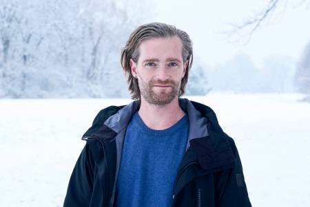 Ein junger Mann mit langem zurückgegelten haaren steht in einer Winterlandschaft