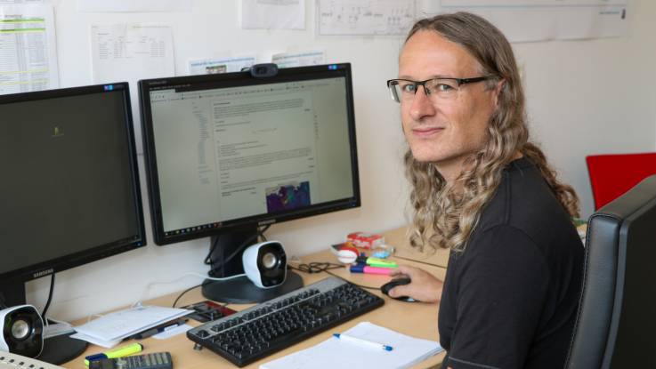 Ein junger Mann mit langen goldblonden Locken sitzt vor zwei großen Bildschirmen und dreht sich zur Kamara um.