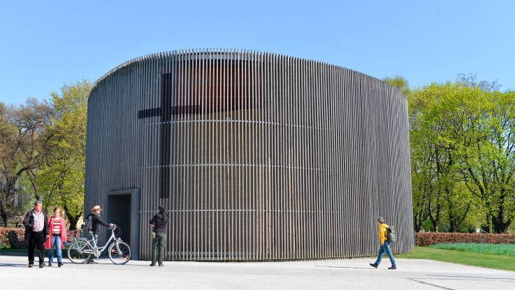 Sachlicher ovaler Bau, der von senkrechten Holzlamellen umgeben ist. An der oval-schmalen Seite ist ein großes christliches Kreuz eingearbeitet.