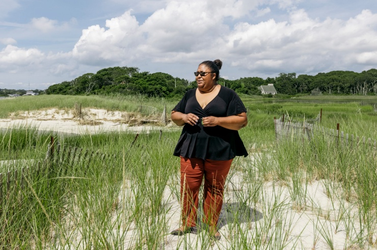 Eine dunkelhäutige Frau steht in einem mit Gras bewachsenen Küstenstreifen und schaut Richtung Meer.