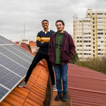 Zwei junge Männer posieren symphatisch lächelnd neben Photovoltaikmodulen auf einem Dach in Lissabon.