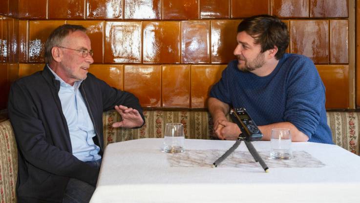 Wolf Dieter Drescher und Sebastian Sladek sitzen vor einem Kachelofen an einem weiß gedeckten Tisch, vor ihnen ein Aufnahmegerät.