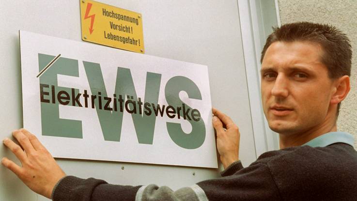 Ein junger Mann mit Bürstenhaarschnitt platziert ein neues EWS-Schild an einer Metalltür und schaut dabei in die Kamera.