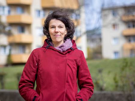 Eine Frau mit dunkelbraunen Haaren und roter Jacke steht vor einem Häuserblock und schaut lächelnd in die Kamera.