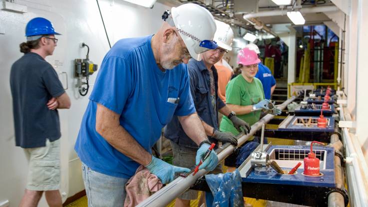 An Bord des Schiffes arbeitet eine Gruppe von Männern und Frauen mit Schutzhelmen bekleidet an einem langen Rohr.