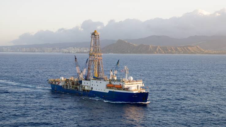 Ein blau-weißes Schiff mit Bohrturm und Kranauslegern fährt einige Kilometer vor einer Küste entlang.