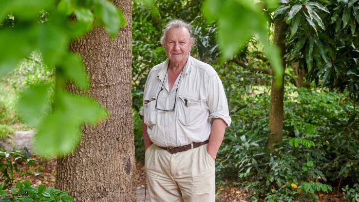 Ein älterer Mann in heller Kleidung steht mit beiden Händen in den Taschen im Wald und blickt in die Kamera.