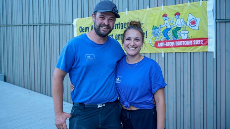 Arm in Arm stehen gutgelaunt eine junge Frau und ein junger Mann, beide in blauen EWS T-Shirts.