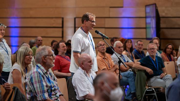 Blick auf das auf Stühlen sitzende Publikum, in dessen Mitte ein Mann in ein Mikrofon spricht.