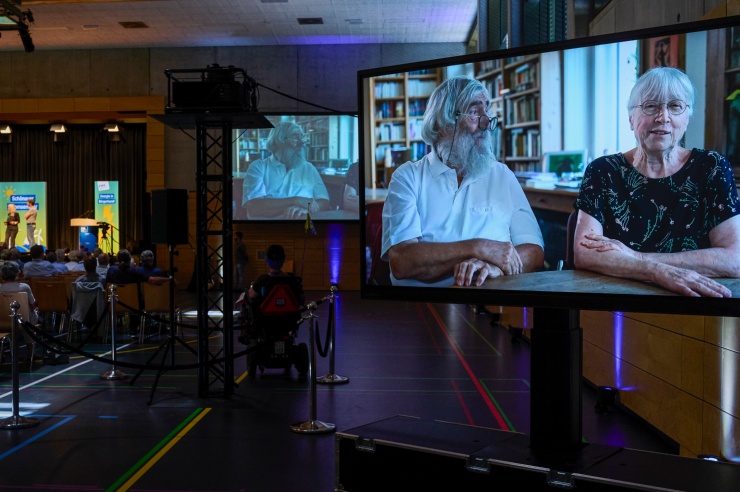 Blick in den Saal mit großen Monitoren, auf denen Ursula und Michael Sladek am Tisch sitzen und in die Kamera sprechen.