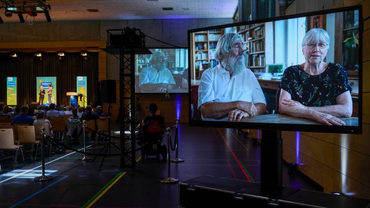 Blick in den Saal mit großen Monitoren, auf denen Ursula und Michael Sladek zu sehen sind, die am Tisch sitzen und in die Kamera sprechen.