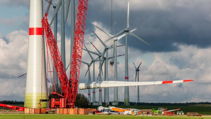 Windkraftanlage im Aufbau: Ein großer roter Kran hebt ein Rotorblatt hoch, der Rest der Baustelle wirkt winzig.