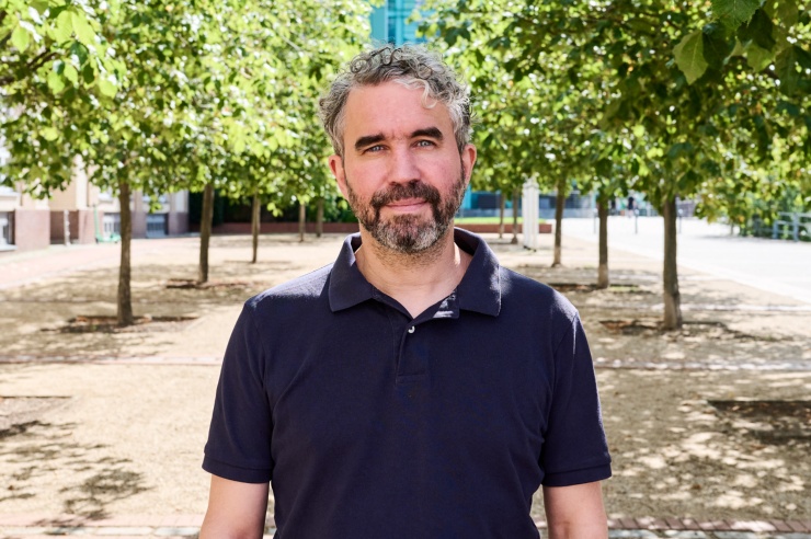 Dennis Tänzler, ein Mann mit grau-melierten Locken und Vollbart vor sommerlichen Bäumen.