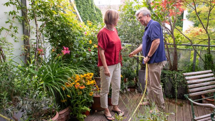 Ein Pärchen bei der Gartenarbeit auf einer Terrasse – der Mann gießt die zahlreichen Pflanzen mit dem Schlauch. 