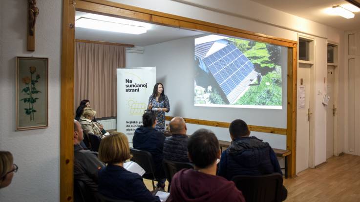 A woman with black hair gives a PowerPoint presentation to some audience members who are taking notes.